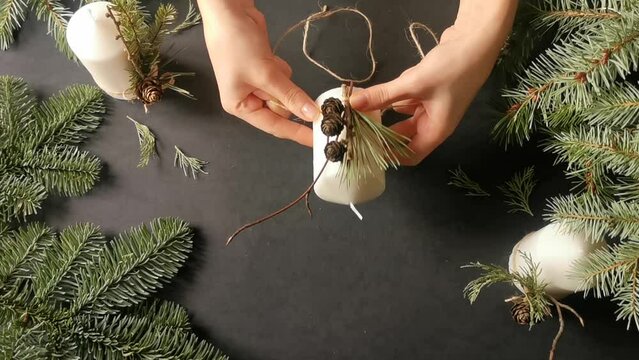 Top view of florist hands making a christmas decor for candles with fir branch, pine cones and thread twine in rustic style. Black background. Eco decor for winter holidays. DIY and handicraft concept