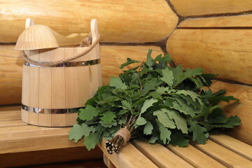 A fresh oak broom and traditional sauna accessories are on a wooden bench in a log corner of bath interior. 