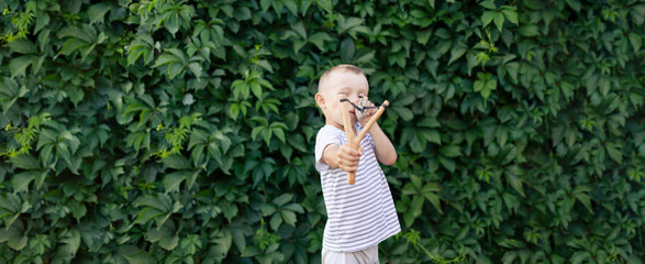 a boy in a T-shirt on the background of a hedge aims a slingshot. banner with space for text