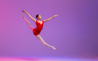 A young ballerina in a red leotard performs a split jump on a lilac background.