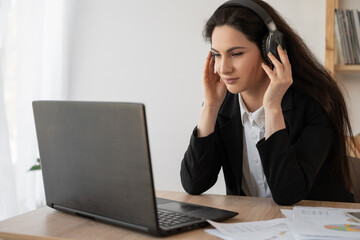 Serious businesswoman in headphones watching webinar on laptop, learning studying computer course, making call, participating online conference