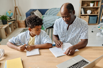 African tutor pointing at copybook and explaining ne material to pupil at table at home schooling