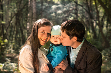 Portrait of a happy young family spending time together in summer nature, on vacation, in nature. Mom and dad hold baby son in their arms and kiss