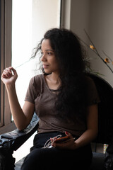 Lifestyle portrait of a curly hair brunette girl sitting in front of a large window. Indian lifestyle