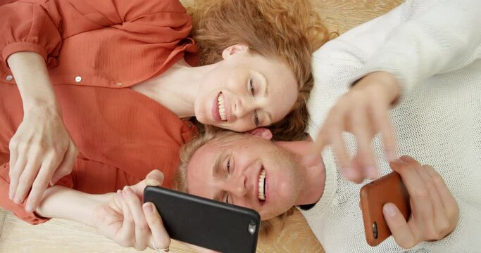 Smiling in love couple on their phone lying on a floor scrolling on the web and social media. Above view of young partners looking at online content. Happy adults looking through technology together