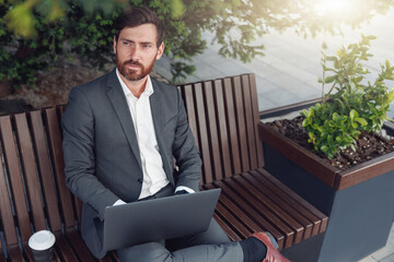 Successful businessman working on laptop at lunchtime sitting on bench near office and looking away