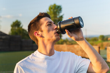 A young man is taking a break during the training to drink water and rest outside in nature during the day