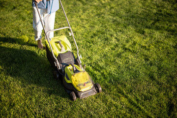 Man mows the lawn with lawn mower, close-up on machine. Concept of modern electric wireless garden equipment - obrazy, fototapety, plakaty