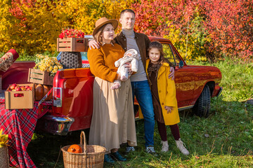 A full-bodied mother with a newborn and little daughter and father stands near a red retro car loaded with boxes of vegetables against the backdrop of rural autumn nature.