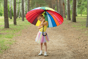 Happy funny girl holding rainbow umbrella. Cute Happy schoolgirl playing in rainy summer park. Kid walking in autumn shower. Outdoor fun by any weather