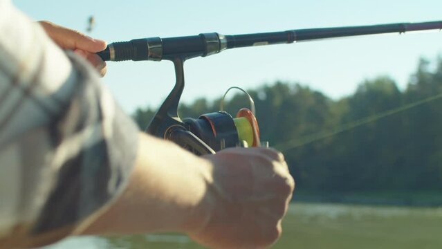 Close-up of African American fisherman holding spin fishing rod, turning fishing reel while angling on pond on summer day.