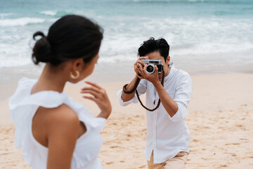 Asian couple enjoying beach vacation holiday. Boyfriend photographer taking a picture of his girlfriend by seaside in summer. A couple wearing a white shirt and dress.