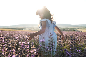 Charming girl in a blue dress and long hair dances in a lavender field in the sunset.