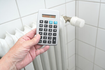 Hand holding a calculator in front of an older heater radiator, energy crises increasing the price of gas and oil, copy space, selected focus