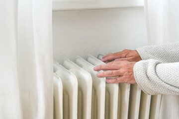 Hands of an elderly woman in woolen clothes feeling low temperature on an old heater, suffering from inflation and rising energy prices, copy space, selected focus - obrazy, fototapety, plakaty
