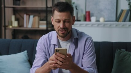 Attractive smiling happy young man use phone sitting on sofa at home. Communication businessman chatting scrolling social media. Close up. Portrait. Slow motion - Powered by Adobe