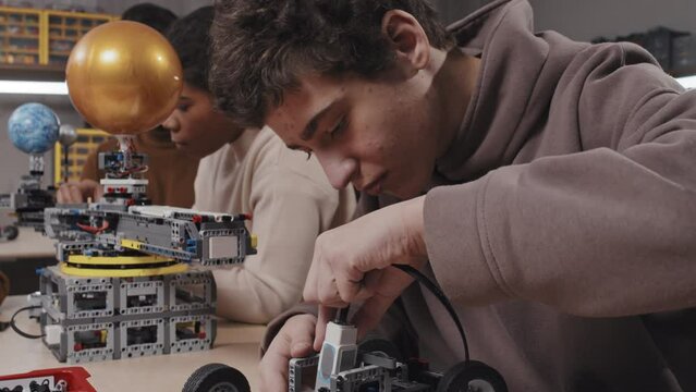Modern teen boy wearing hoodie sitting at table building car model during robotics class at school