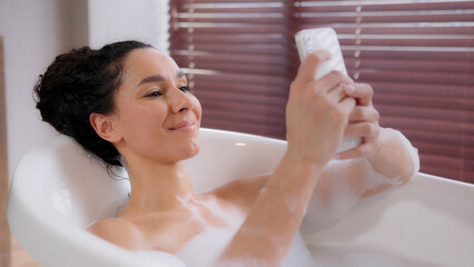 Young happy hispanic woman lying in hot comfortable foam luxurious bath relaxed girl typing sms on...