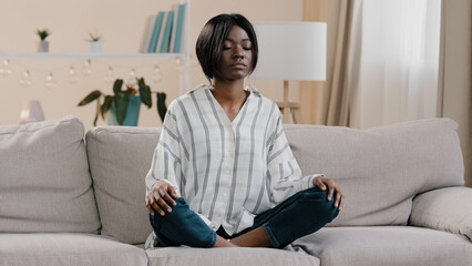 Young focused african american woman sitting in lotus position with closed eyes healthy calm girl relaxing on sofa calming down taking deep breath meditating relaxing at home getting rid of stress