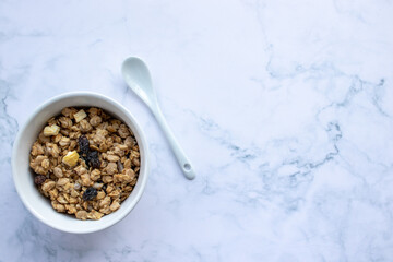 Bowl of homemade granola cereal with ceramic spoon on white marble table background. Top view, flat...
