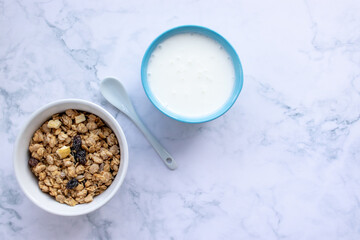 Bowl of homemade granola cereal with greek yogurt and ceramic spoon on white marble table background. Top view, flat lay, copy space. Healthy eating concept. 