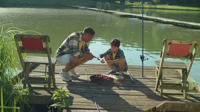 Skillful handsome African fisherman and adorable preadolescent Arab boy sitting on wooden pier, talking about fishing lures and spinning baits for fishes while family preparing for fishing by lake.