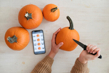 Young woman preparing to carve Jack O Lantern from a pumpkin using stencils from the internet. Female making Halloween decorations with tutorial on the phone. Background, copy space.