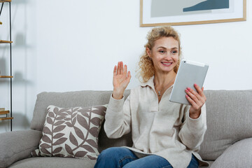 Obraz premium Portrait of adult blonde woman wearing mom jeans sitting on the couch holding a tablet device and smiling. Joyful female having fun video call at home. Background, copy space, close up.