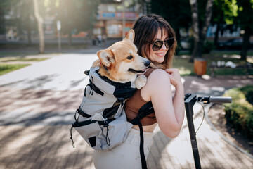 Happy smiling woman traveler is riding her electro scooter in city parkland with dog Welsh Corgi Pembroke in a special backpack - 522252241