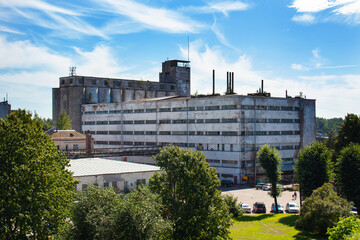 Old grain elevator and gray building among green trees under blue sky