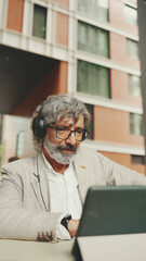 Mature businessman with beard in eyeglasses and headphones, sits in an outdoor cafe and works using tablet