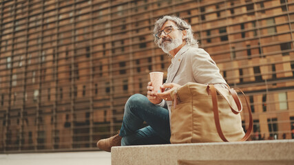 Mature businessman with beard in eyeglasses wearing gray jacket holding takeaway coffee. Middle aged manager enjoying morning coffee while sitting outside the office