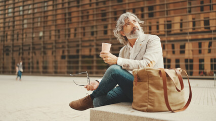 Mature businessman with beard in eyeglasses wearing gray jacket holding takeaway coffee. Middle...
