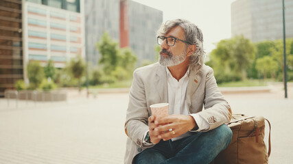 Mature businessman with beard in eyeglasses wearing gray jacket holding takeaway coffee. Middle...