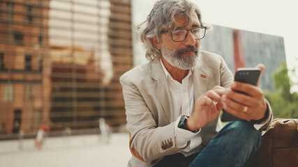 Mature businessman with beard in eyeglasses wearing gray jacket is using cell phone. Middle aged...