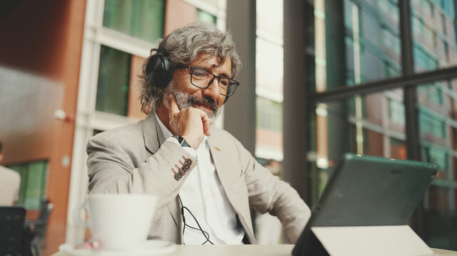 Mature businessman with beard in eyeglasses and headphones, sits in an outdoor cafe and works using tablet