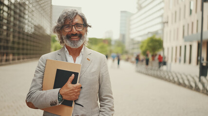 Smiling mature businessman with beard in eyeglasses wearing gray jacket looks around and walks down...