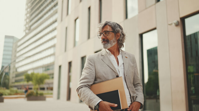 Fototapeta Smiling mature businessman with beard in eyeglasses wearing gray jacket looks around and walks down the street past modern buildings