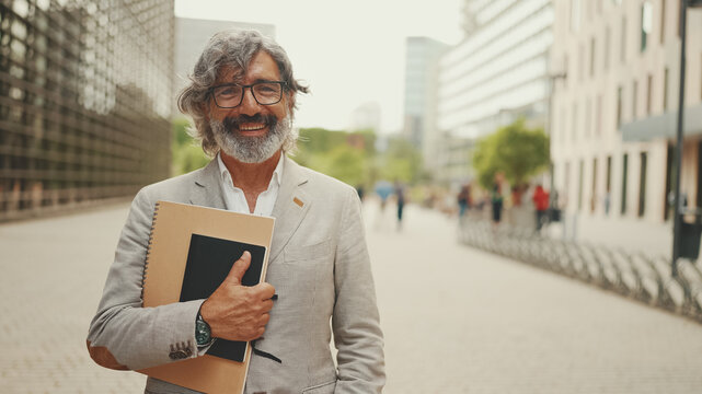 Fototapeta Smiling mature businessman with beard in eyeglasses wearing gray jacket looks around and walks down the street past modern buildings