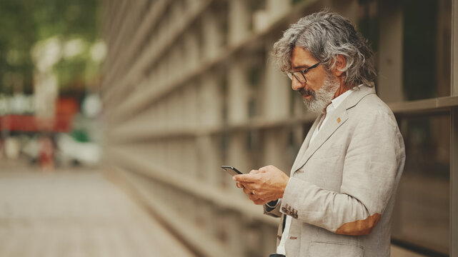 Fototapeta Profile mature businessman with beard in eyeglasses wearing gray jacket looking up at street signs and map trying to find her way using cellphone