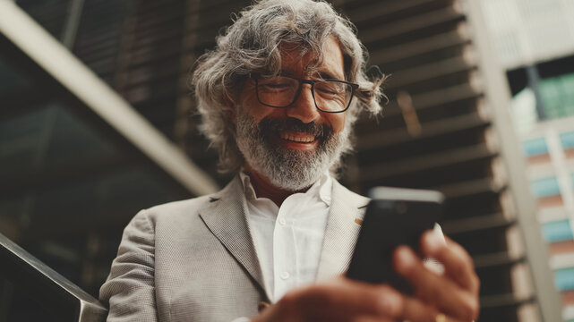 Mature Businessman With Beard In Glasses Stands On The Street At The Business Center Background. Middle Aged Manager Texting Message Scrolling Tapping To Smartphone. Joy From Good Deal