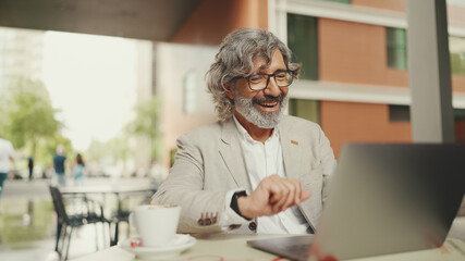 Mature businessman with beard in eyeglasses wearing gray jacket sits drinks coffee in cafe. Middle...