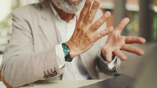 Close-up of the hands of mature businessman having video conference, video call on laptop pc computer sit at cafe outdoor. Middle aged manager massage hands, gesture
