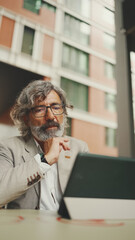 Fototapeta premium Mature businessman with beard in eyeglasses wearing gray jacket, working on tablet, sitting in an outdoor cafe. Successful man sitting at table in cafe in nature Mobile office freelancer