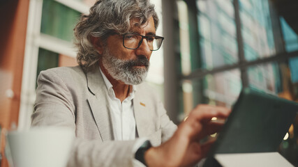 Naklejka na ściany i meble Mature businessman with beard in eyeglasses wearing gray jacket, working on tablet, sitting in an outdoor cafe. Successful man sitting at table in cafe in nature Mobile office freelancer