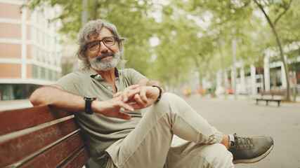 Obraz premium Thoughtful middle-aged man with gray hair and beard wearing casual clothes sits on bench. Mature gentleman in eyeglasses turns his head and looks into the camera