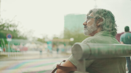 Thoughtful, middle-aged man with gray hair and beard, wearing casual clothes, sits at public transport stop. Mature gentleman in eyeglasses with bag is waiting for the tram at the tram station