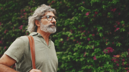 Friendly middle-aged man with gray hair and beard, walks next to wall with purple flowers blooming in the background