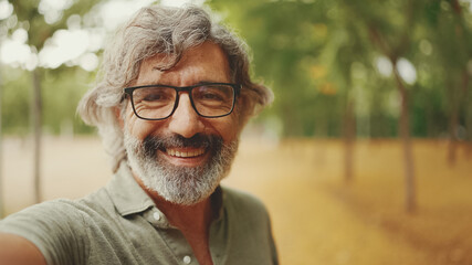 Clouse-up, friendly middle-aged man with gray hair and beard wearing casual clothes looks at the camera. Mature gentleman in eyeglasses smiles at the camera