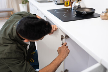 Blurred craftsman with screwdriver fixing metal hinge in kitchen cabinet
